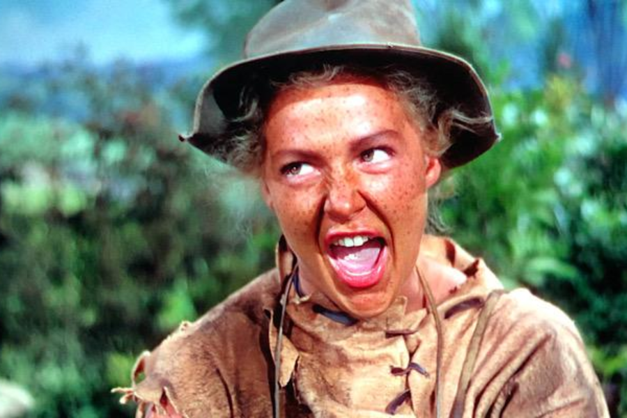 Close-up scene from a film showing a freckled woman in a tattered, tan frontier outfit and floppy hat, shouting or singing with her mouth wide open. Her exaggerated expression and costume suggest a comedic or theatrical portrayal of a young Annie Oakley.
