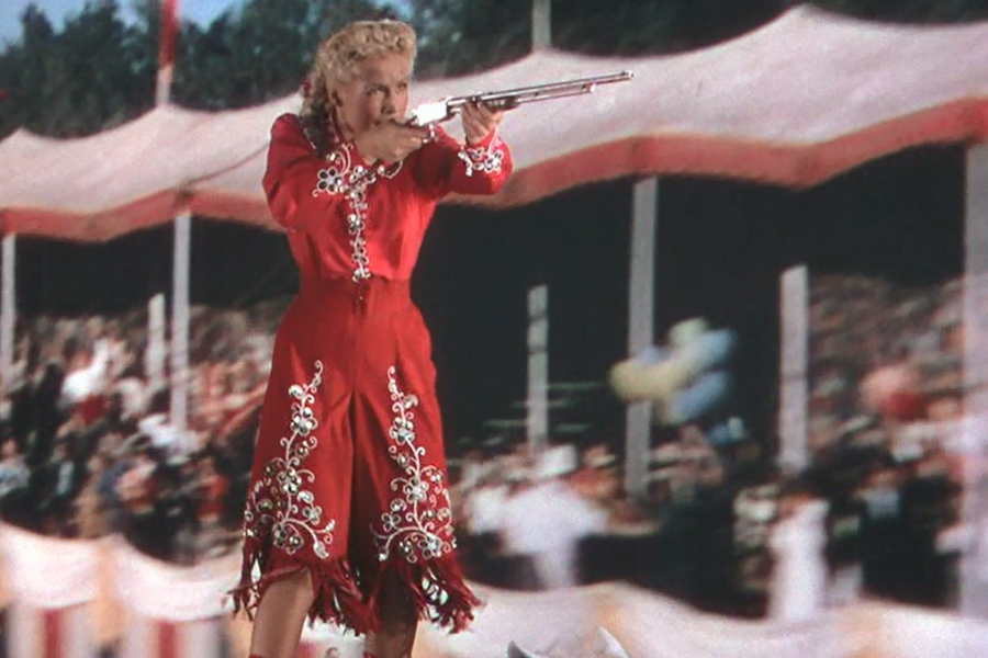 Colorful film scene of a blonde woman in a bright red Western outfit with white embroidery, aiming a rifle with precision. She appears to be performing in front of a large, cheering crowd under striped circus-style tents, capturing the flair of a Wild West show.