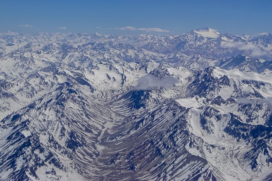 Aerial view of the Andes Mountains covered in snow, with jagged peaks stretching across the horizon. Deep valleys and ridges weave through the range under a bright, cloud-dotted blue sky.
