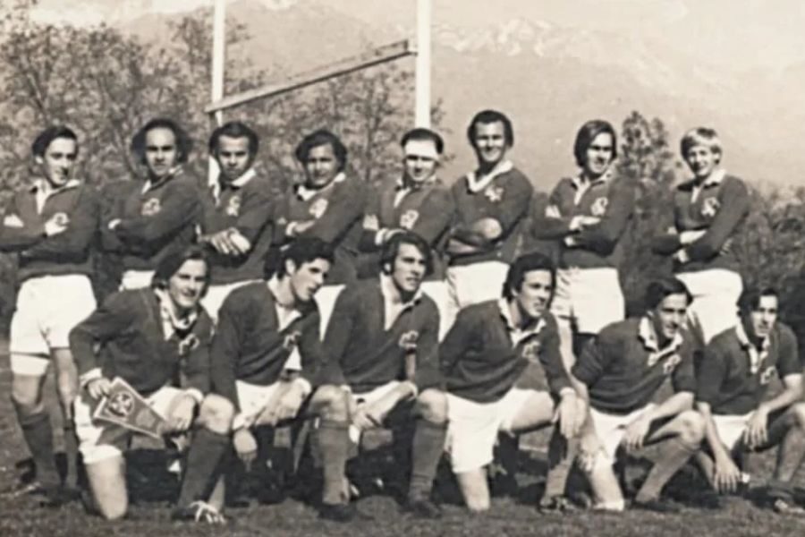 A black-and-white photo of a young men's rugby team, likely from the 1970s. The players wear matching jerseys with a crest, posing proudly on a field with trees and faint mountains in the background.