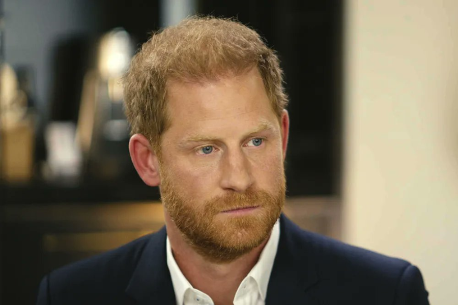 A man with reddish-blond hair and a neatly trimmed beard sits indoors in a formal setting, wearing a navy blazer and white shirt. He looks slightly off-camera with a serious, contemplative expression.