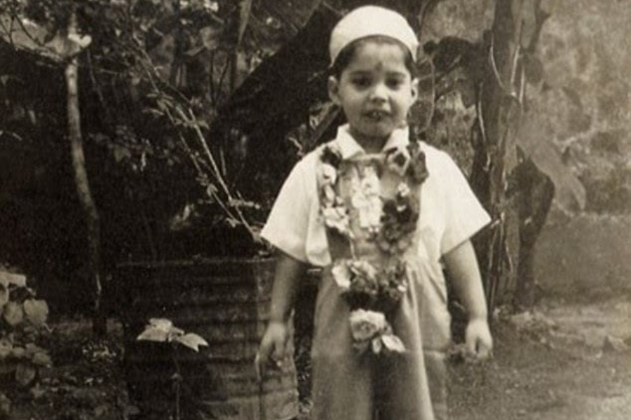 A young child dressed in short overalls, a light-colored shirt, and a cap stands outdoors, wearing a garland made of flowers. The photo is in black and white, with a rustic background of leafy plants and a tin barrel, evoking a vintage atmosphere.