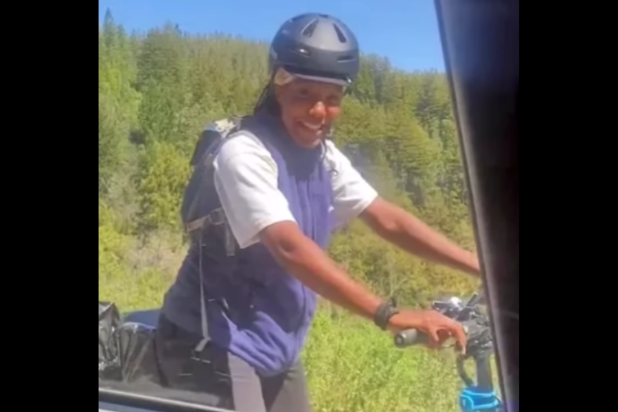 A smiling cyclist wearing a helmet, short-sleeve shirt, and vest poses next to their bike on a sunny trail lined with lush green trees. The person appears to be speaking to someone inside a vehicle, partially visible on the right edge of the image.