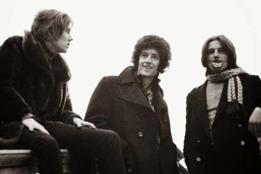 Vintage black-and-white photo of three young people dressed in winter coats, standing or sitting casually against a bright sky backdrop. The person on the left looks toward the others while sitting on a ledge, the center figure smiles warmly, and the one on the right wears a striped scarf and grins playfully with fake novelty teeth.