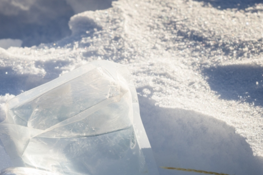 A translucent plastic water container partially filled with liquid rests on a sunlit patch of snow, with sparkling ice crystals reflecting the light around it. The container appears flexible, likely used for outdoor or alpine activities.