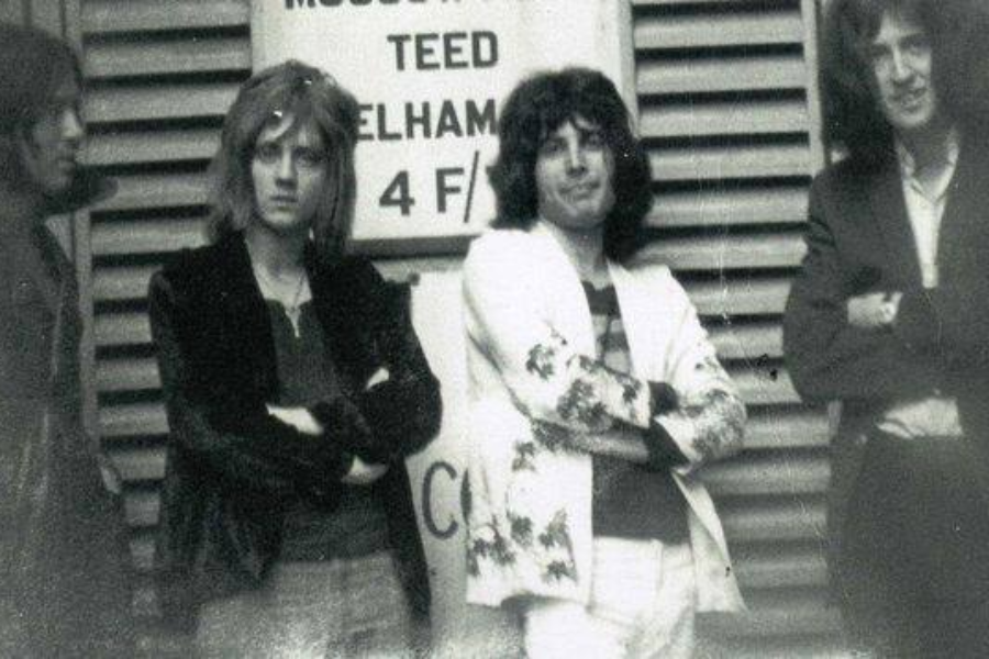 Grainy black-and-white photo of four young men standing casually in front of a building with louvered panels and a sign that reads “MUSCOTEEED PELHAM 4 F.” The group, dressed in rock-style clothing with long hair and confident postures, includes a man in a white jacket with floral patterns who stands out at the center.