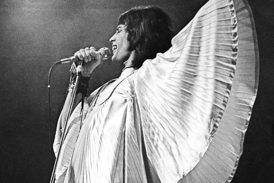 Black-and-white image of a flamboyant male performer singing passionately into a microphone on stage. He wears a dramatic, pleated cape-like outfit with wide, winged sleeves extended outward, capturing a dynamic moment mid-performance under concert lighting.