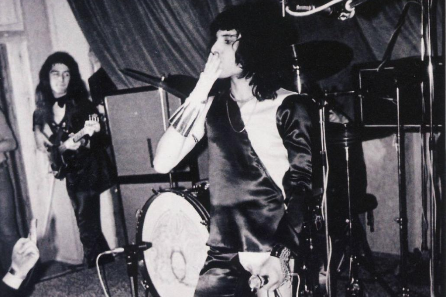 Black-and-white photo of a live concert scene showing a dramatic moment as a lead singer, dressed in a sleek satin outfit with metallic arm cuffs, blows a kiss to the audience. A guitarist stands in the background near a drum kit with Queen's crest visible, indicating an early performance by the band Queen.