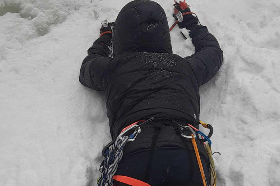 A climber wearing a black insulated jacket and a harness equipped with carabiners and climbing gear ascends a snowy or icy slope using ice tools. The person is positioned belly-down on the snow, fully engaged in a technical winter ascent.