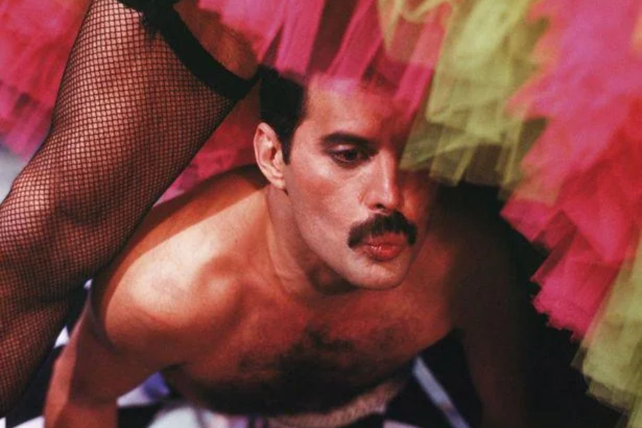 Vibrant close-up of a shirtless man with a mustache, crouched beneath a flurry of colorful tulle skirts in pink, yellow, and green. A fishnet-clad leg is visible in the foreground, adding a theatrical and playful tone to the scene, with the man’s expression appearing focused or contemplative.
