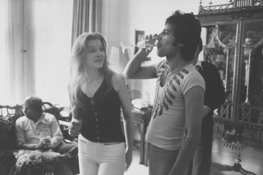 Black-and-white photo of a relaxed social gathering in an elegantly furnished room. A man in a fitted, graphic t-shirt (Freddie Mercury) drinks from a champagne glass while standing next to a smiling woman with long blonde hair. Other guests lounge in the background, adding to the laid-back, intimate atmosphere.