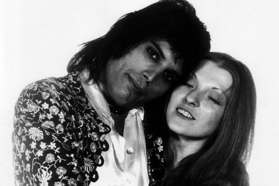 Black-and-white portrait of a man and woman embracing closely, both appearing serene and affectionate. The man, dressed in a satin shirt and ornate floral jacket, leans his head gently against the woman, who has long hair and a soft, content expression with her eyes partially closed.