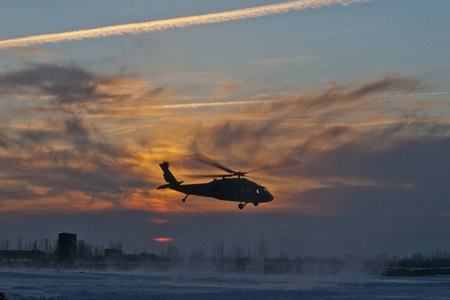 A military helicopter is silhouetted against a dramatic sunset sky, hovering above a snowy landscape with swirling snow clouds below it. Contrails and streaks of clouds stretch across the colorful evening sky.