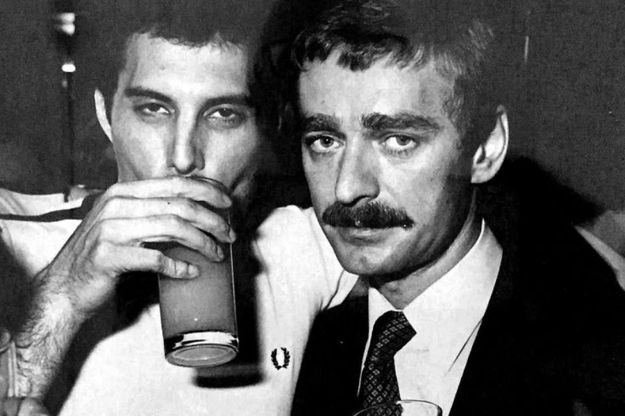 Black-and-white photo of two men closely posed at what appears to be a bar or social gathering. On the left, a man in a casual polo shirt drinks from a glass while looking directly at the camera. On the right, a man with a mustache in a suit and tie also faces the camera, his expression composed and slightly solemn.