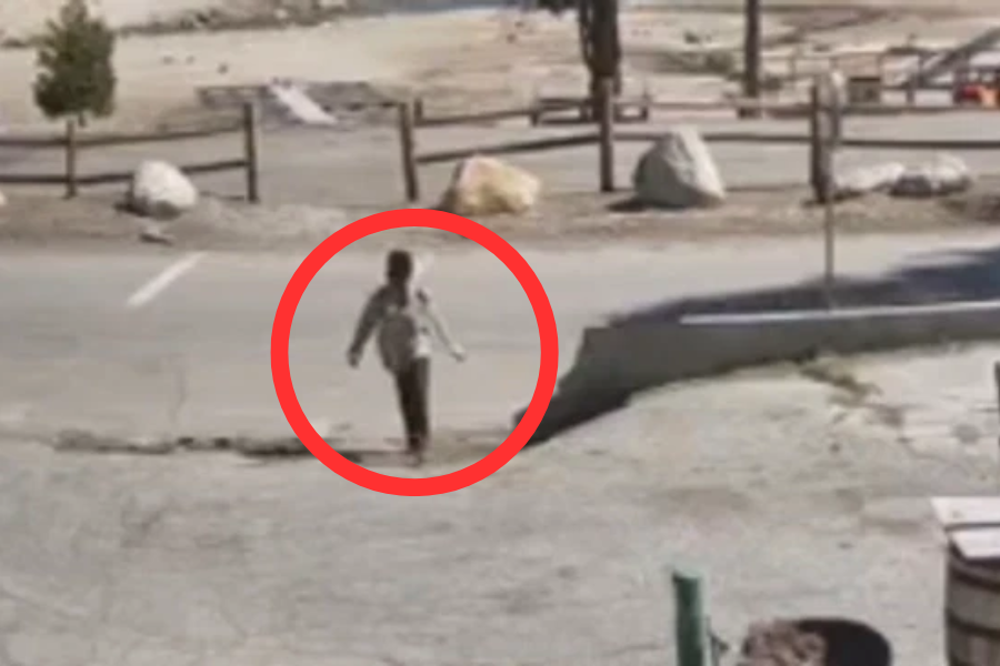 A surveillance-style image shows a person walking alone across a mostly empty paved area, possibly a parking lot, with a red circle digitally added to highlight them. The background includes large rocks, wooden fences, and a sandy or dry landscape.