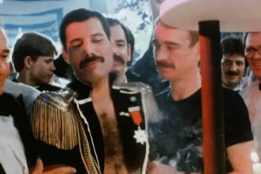 Color photo of Freddie Mercury dressed in a theatrical black military-style jacket with gold epaulettes and medals, surrounded by a joyful group of people at a party. Standing beside him is Jim Hutton in a black t-shirt, with both men smiling amidst the festive atmosphere and crowd of onlookers in semi-formal attire.