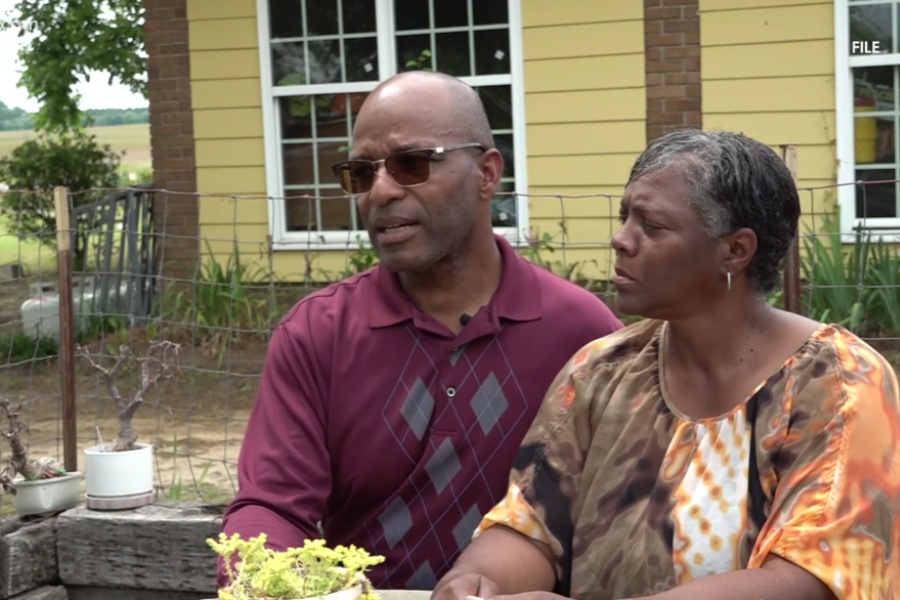 A man and woman sit together outside a yellow house, appearing serious and emotional. The man wears sunglasses and a maroon shirt with an argyle pattern, while the woman, dressed in a multicolored blouse, looks at him with concern. Potted plants and a rural backdrop suggest a country setting.