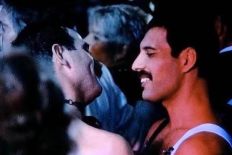 Color photograph of Freddie Mercury smiling warmly at another man during a crowded event or party. Both men appear joyful and close, with Mercury wearing a white tank top and a signature mustache, while the other man wears a choker necklace. The background is filled with blurred guests, adding to the intimate, celebratory mood.