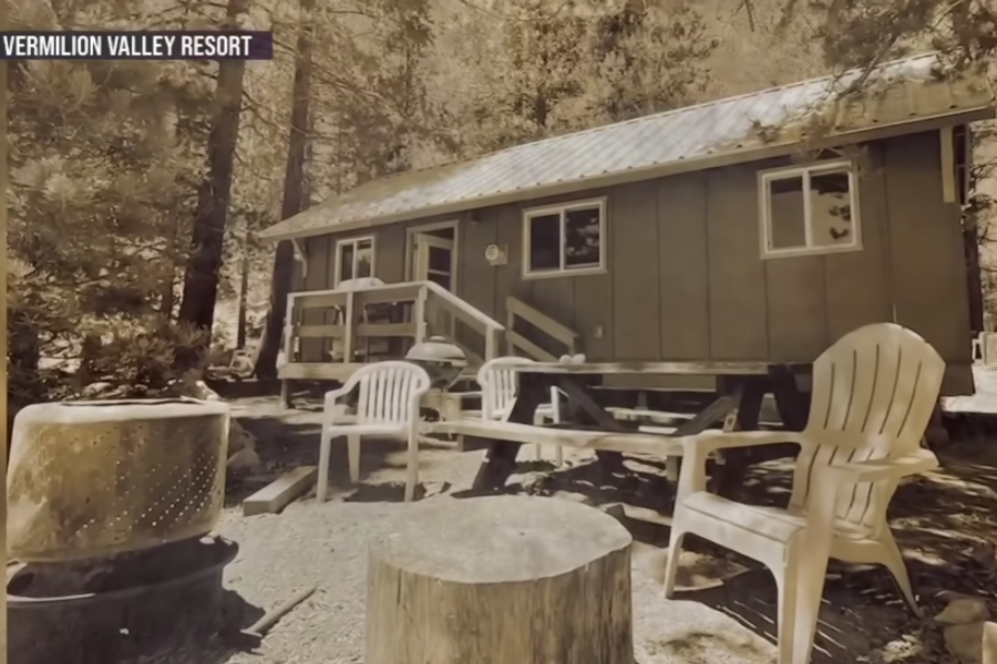 A sepia-toned photo of a rustic cabin at Vermilion Valley Resort, surrounded by tall pine trees. In front of the cabin, there are plastic chairs, a picnic table, a fire pit made from a repurposed metal drum, and a tree stump, creating a cozy outdoor sitting area.