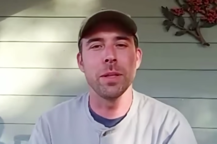 A man wearing a baseball cap and light-colored shirt sits in front of a light green paneled wall, speaking or being interviewed. There’s a decorative branch with red berries mounted on the wall behind him.