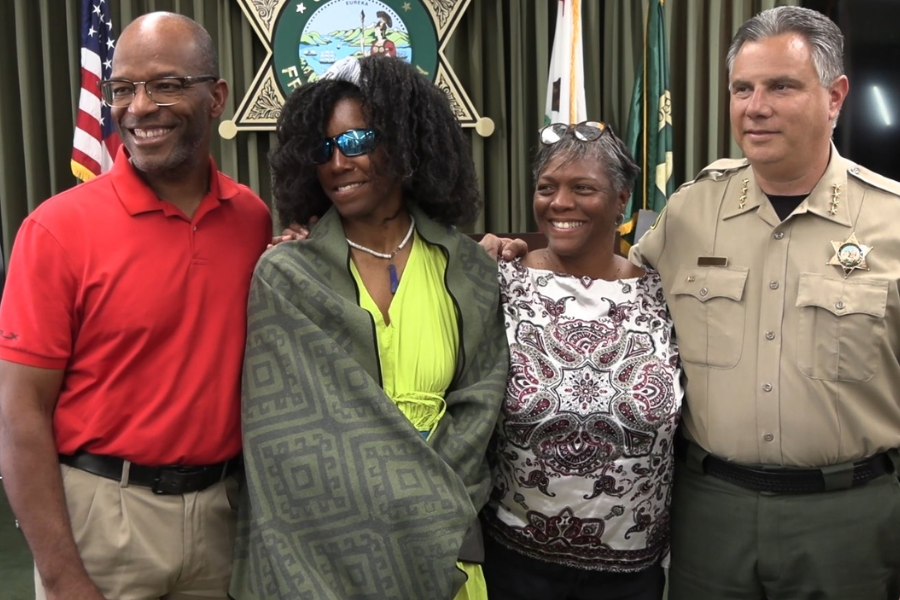 Four people stand smiling for a photo in front of a sheriff's department emblem and flags. From left to right: a man in a red polo shirt, a woman in a bright green dress with a green patterned blanket draped over her shoulders and wearing sunglasses, a woman in a white patterned blouse, and a uniformed sheriff.