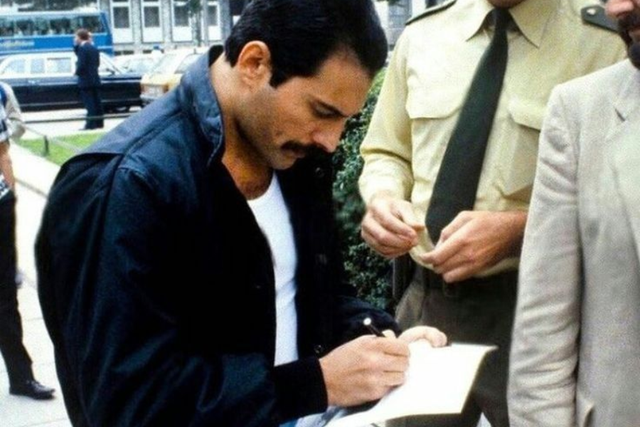 Candid color photo of Freddie Mercury outdoors, wearing a black jacket over a white shirt, as he signs an autograph for a fan. He’s focused on writing, with uniformed individuals and others standing nearby, likely during a public appearance or arrival.