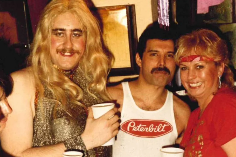 Freddie Mercury stands smiling between two partygoers at a lively gathering. He wears a white tank top with a red "Peterbilt" logo, while the person on the left is dressed in a sparkly outfit with a long blonde wig and the person on the right sports a red top and matching headband. All three hold cups, suggesting a fun and relaxed celebration.