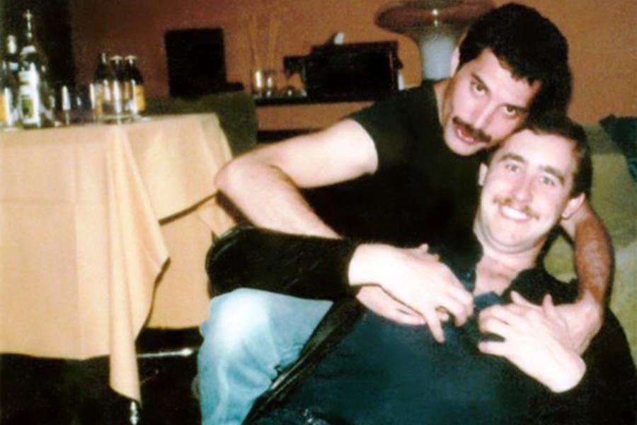 Candid, warmly lit photo of Freddie Mercury embracing Jim Hutton from behind in a playful and affectionate moment. Both are smiling, with Mercury wearing a black shirt and wrist cuff, and Hutton leaning back comfortably into his arms. A table with bottles and a softly lit room create an intimate, relaxed setting.