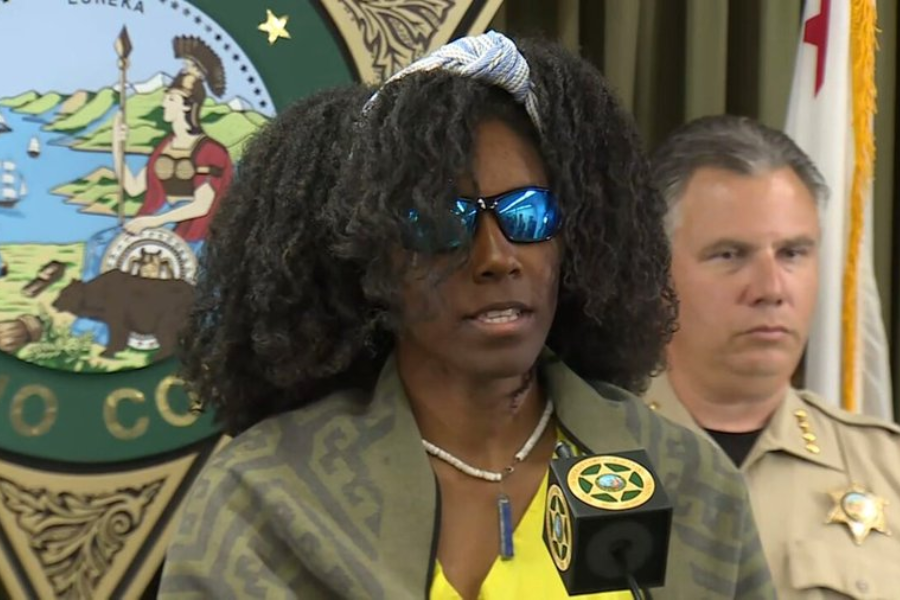 A woman wearing sunglasses, a headband, and a green patterned wrap speaks at a press conference podium with a law enforcement badge symbol on the microphone. A uniformed sheriff stands behind her, and the California state seal is visible in the background.