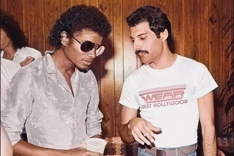 Iconic photo of Michael Jackson and Freddie Mercury engaged in quiet conversation. Michael Jackson wears aviator sunglasses and a light floral button-up shirt, while Freddie Mercury sports a white “WEAR West Hollywood” t-shirt and holds a cigarette. The wood-paneled background suggests a casual, private indoor setting.