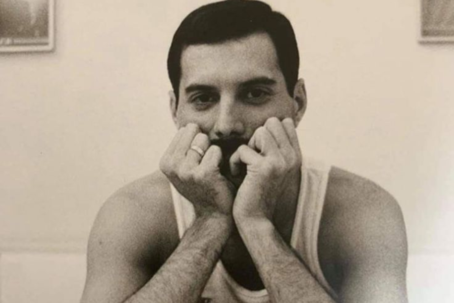 Black-and-white portrait of Freddie Mercury resting his chin on his fists, elbows on the table, gazing directly at the camera with a soft, thoughtful expression. He wears a sleeveless shirt, and two rings are visible on his fingers, creating an intimate and quietly powerful moment.