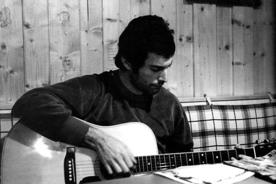 Black-and-white photograph of Freddie Mercury sitting in a cozy, wood-paneled room, deeply focused while playing an acoustic guitar. He wears a sweatshirt and sits on a plaid-upholstered bench, with papers or magazines spread out in front of him, capturing a quiet and creative moment.