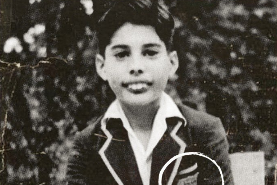 Vintage black-and-white portrait of a young boy seated outdoors, wearing a dark blazer with light piping and a crest on the pocket, partially highlighted by a white circle. The boy looks directly at the camera with a composed expression, set against a leafy background.