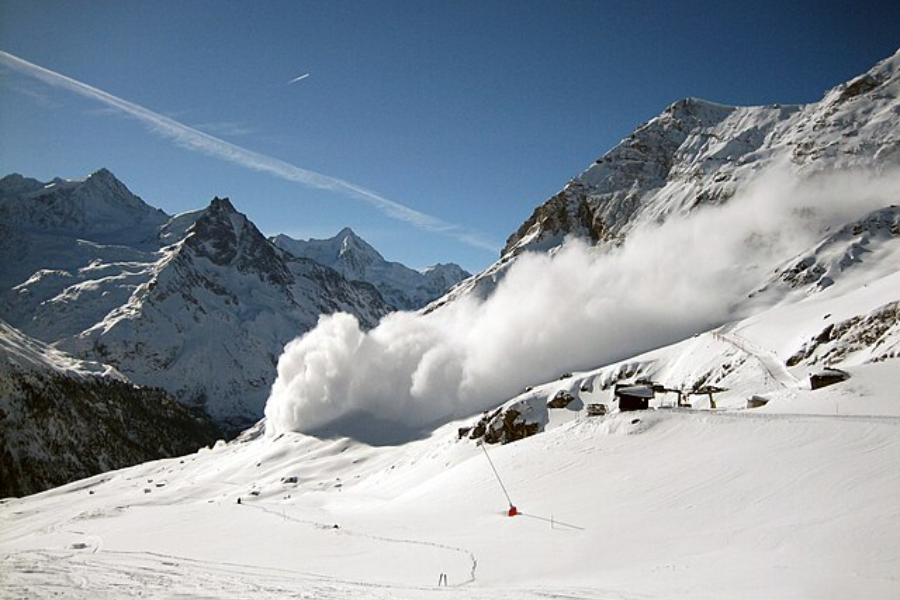 A powerful avalanche cascades down a snowy mountain slope in a high alpine region under a clear blue sky, with a few ski lodges and lift structures visible in the foreground. The snow cloud from the avalanche spreads widely, contrasting against the jagged, snow-covered peaks in the distance.