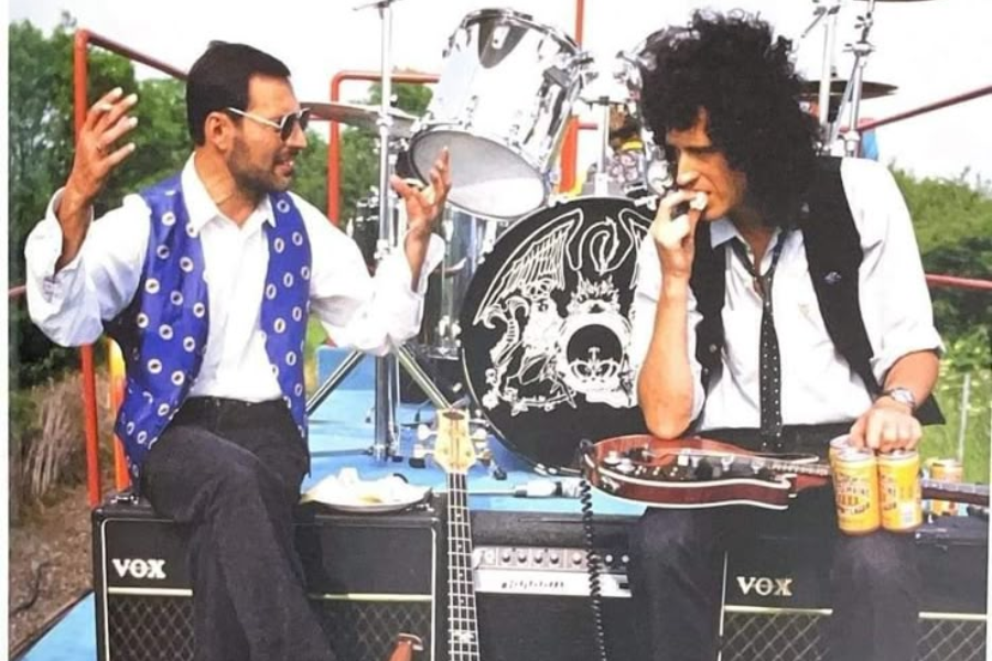 Freddie Mercury and Brian May sit on VOX amplifiers in front of a Queen-branded drum kit outdoors. Freddie, wearing a vibrant blue vest and sunglasses, gestures animatedly while talking. Brian, dressed in a black vest and holding a guitar, listens thoughtfully with a can of beer in front of him. The scene feels like a casual moment during a break in rehearsal or filming.