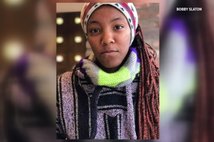 A young woman with reddish braided hair looks calmly at the camera, wearing a multicolored knit scarf, a striped sweater, and a cozy winter hat. The background is softly blurred, with a warm indoor setting and lights behind her.