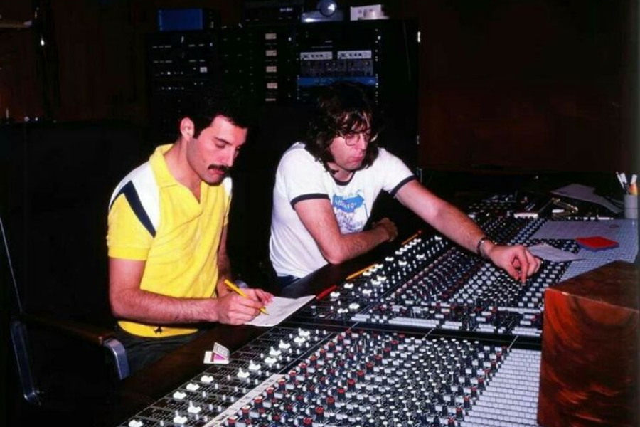 Freddie Mercury, wearing a yellow polo shirt, sits in a recording studio beside a sound engineer at a large mixing console. Freddie writes on a notepad while the engineer adjusts controls, surrounded by rows of dials, sliders, and equipment. The scene captures an intimate, behind-the-scenes moment of music production.