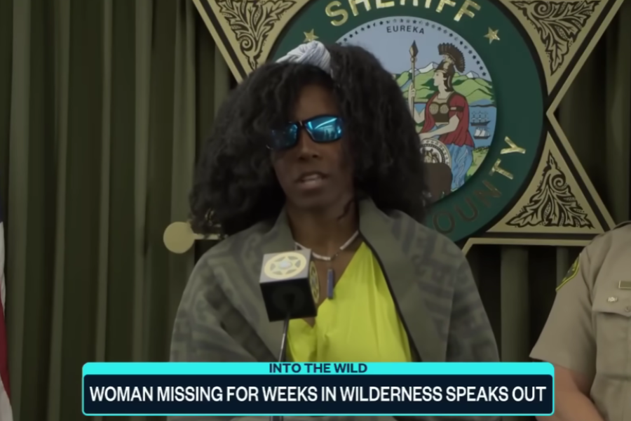 A woman wearing sunglasses and a green patterned shawl speaks at a sheriff's department press conference. Behind her is a large badge emblem and the California state seal, with the news caption below reading: “INTO THE WILD – WOMAN MISSING FOR WEEKS IN WILDERNESS SPEAKS OUT.”