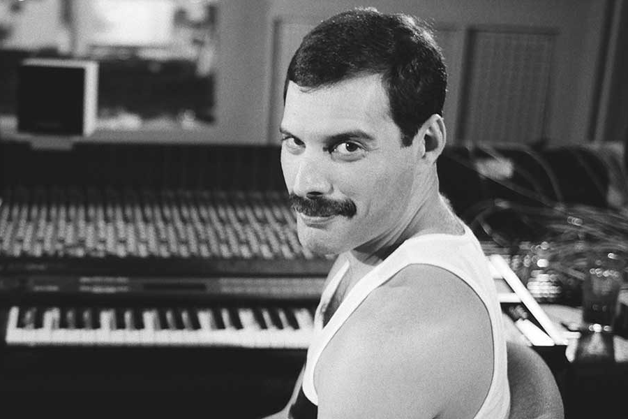 Black-and-white photo of Freddie Mercury seated at a mixing console and keyboard in a recording studio. He wears a white tank top and turns over his shoulder to smile directly at the camera, exuding warmth and quiet confidence amidst the creative workspace.
