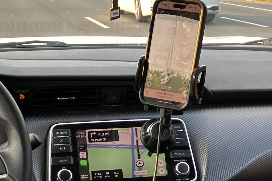 The dashboard of a car showing both an in-dash GPS system and a mounted smartphone with navigation apps directing to W Pratt St. The screen indicates 4.3 miles remaining and an estimated arrival time of 7:22. A road with other vehicles is visible through the windshield.