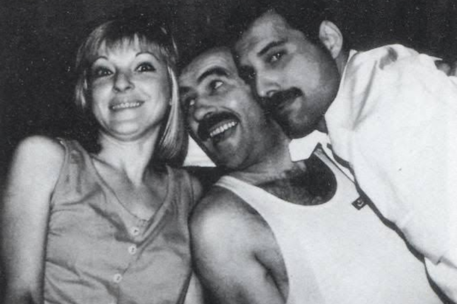 Black-and-white candid photo of Freddie Mercury warmly embracing his smiling parents, Jer and Bomi Bulsara. Freddie leans in close with a soft smile, while his father beams and his mother looks cheerful and surprised, capturing a joyful and intimate family moment.