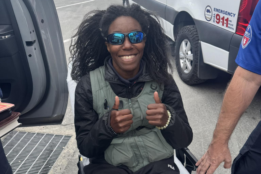 A smiling woman wearing reflective sunglasses and a green vest gives two thumbs up while seated on a stretcher, surrounded by emergency responders. She is outdoors near a 911 emergency vehicle, with a van door open beside her.