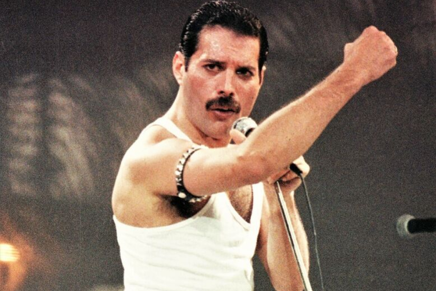 Iconic color photo of Freddie Mercury mid-performance at Live Aid in 1985. Wearing a white tank top and silver armband, he strikes a powerful pose with a raised fist while gripping a microphone, exuding confidence and charisma under dramatic stage lighting.
