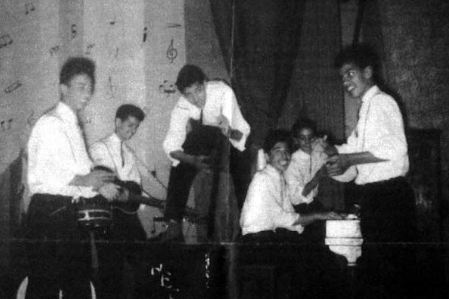 Black-and-white photograph of a lively school band performing on stage, with six young men dressed in white shirts and dark trousers. The group includes musicians playing drums, guitar, upright bass, and piano, with hand-drawn musical notes decorating the backdrop, evoking a joyful, retro concert atmosphere.