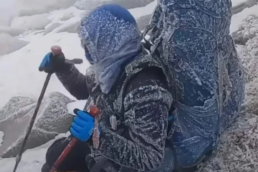 A mountaineer covered in frost and snow sits on a rocky, icy slope holding trekking poles, wearing blue gloves and a large frozen backpack. The extreme cold is evident from the thick ice buildup on their clothing and gear in the harsh alpine environment.