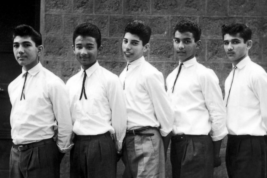 Black-and-white photo of five young men standing in a line outdoors against a brick wall, all dressed in white shirts, dark trousers, and bolo-style neckties. They pose confidently with hands behind their backs, exuding a 1950s or early 1960s student or band look.