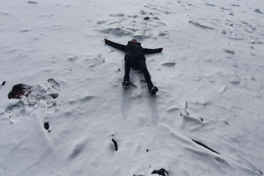 A person dressed in black winter clothing lies on their back in the snow with arms and legs spread out, possibly making a snow angel or simply resting. The snowy ground is lightly trampled with footprints and patches of exposed earth.