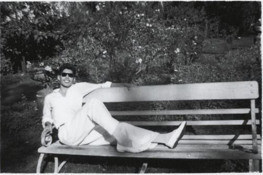 Black-and-white photo of a young man lounging stylishly on a wooden bench in a sunny garden, wearing sunglasses and a light-colored outfit. He reclines with one arm draped over the bench back and legs stretched out, smiling confidently amid blooming bushes and trees.