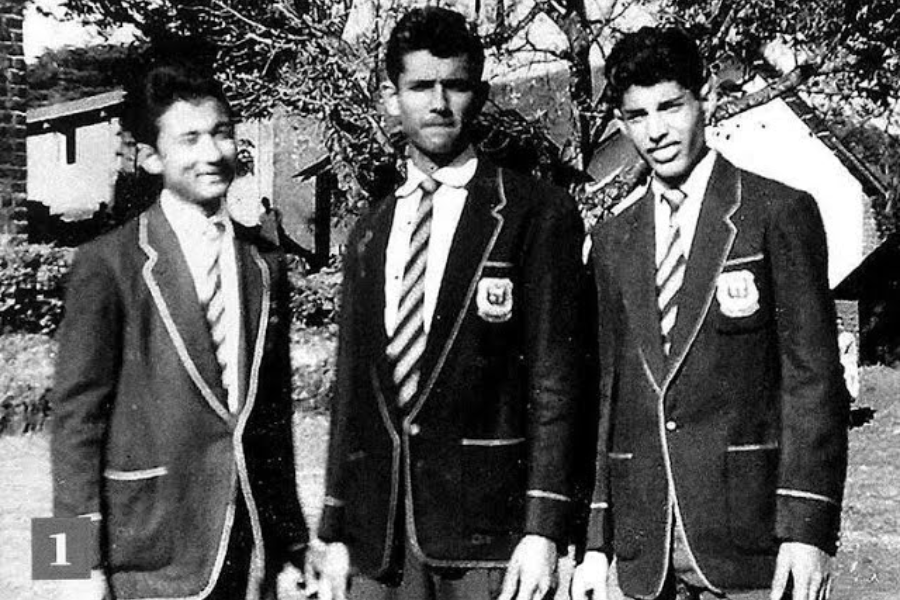 Black-and-white photograph of three young men standing outdoors in matching school blazers with piping and crest patches, paired with striped ties and dress shirts. They pose formally with subtle smiles, likely representing a team or academic group, with school buildings and trees in the background.
