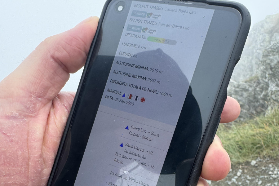 A close-up of a person's hand holding a smartphone displaying hiking trail information in Romanian, including distance (6 km), estimated duration (6 hours), elevation range (2019 m to 2507 m), and route markings. The screen also shows a trail route from Cabana Bâlea Lac to Parcarea Bâlea Lac with a difficulty rating of medium (6/10) and a date of September 5, 2020.